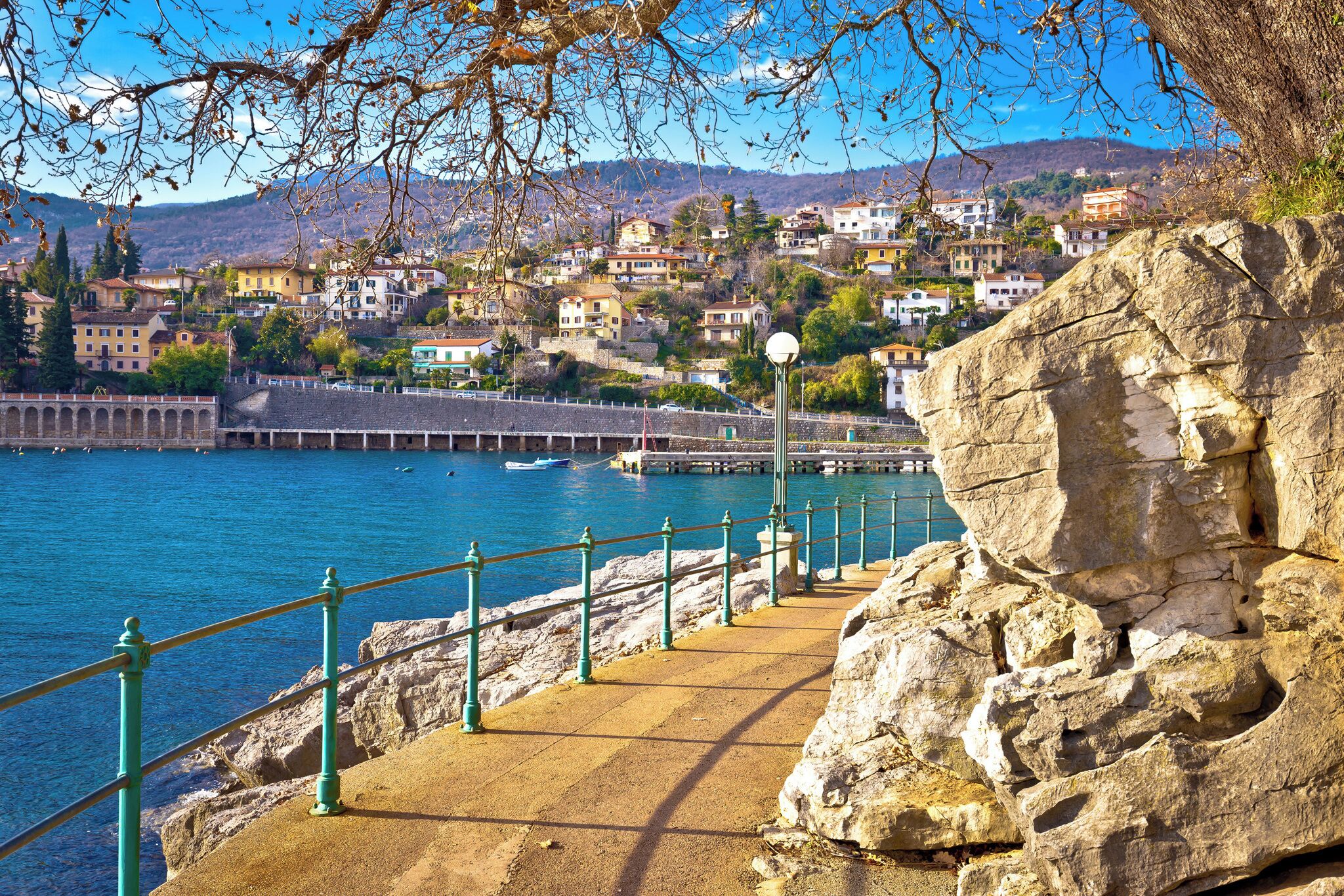 A coastal town with a stone staircase leading down to the water, a rocky outcrop, and a view of buildings in the background.