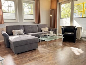 Ein modernes Wohnzimmer mit einem grauen Ecksofa, einem Glastisch, einem schwarzen Ledersessel und einer Stehlampe.