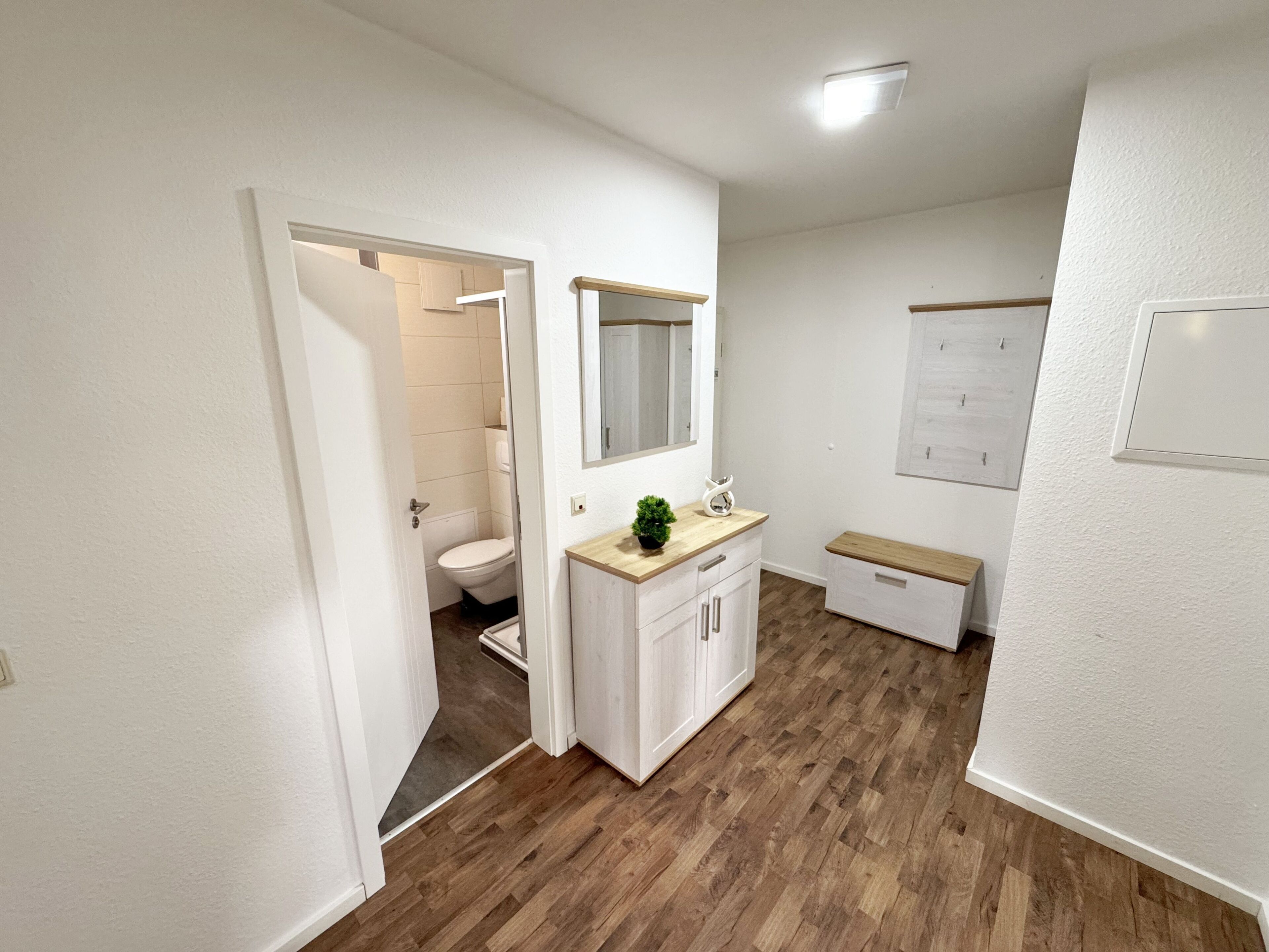 Un baño moderno con suelo de madera, muebles blancos, un espejo sobre el lavabo y un inodoro visible a través de una puerta abierta.