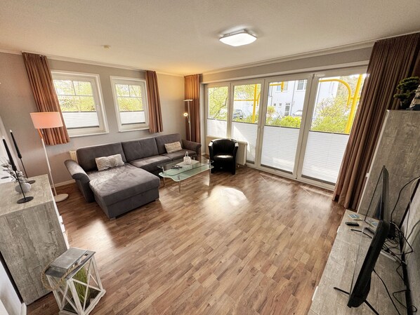 A modern living room with a grey sectional sofa, a glass coffee table, and a black leather chair. There are large windows with curtains, a floor lamp, and a TV stand with a flat-screen TV.