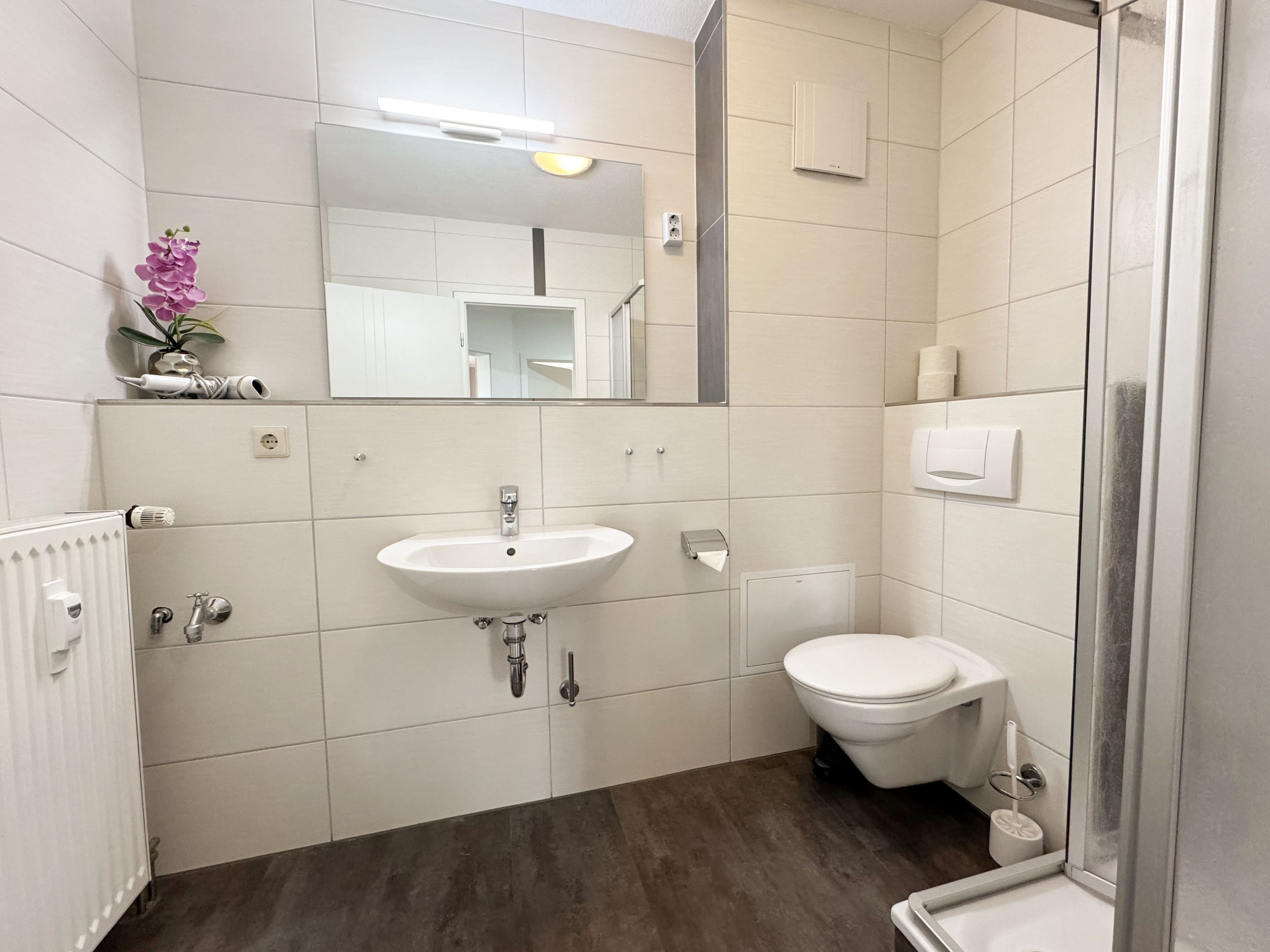 Ein modernes Badezimmer mit Waschbecken, Toilette und Dusche. Über dem Waschbecken hängt ein Spiegel, auf der Ablage steht eine kleine Pflanze.