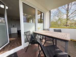 An outdoor patio with a wooden table and chairs, a sliding glass door leading to a kitchen, and a view of trees and a fence.