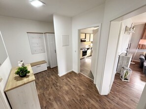 A modern interior with a wooden floor, white walls, and a kitchen visible through an open door.
