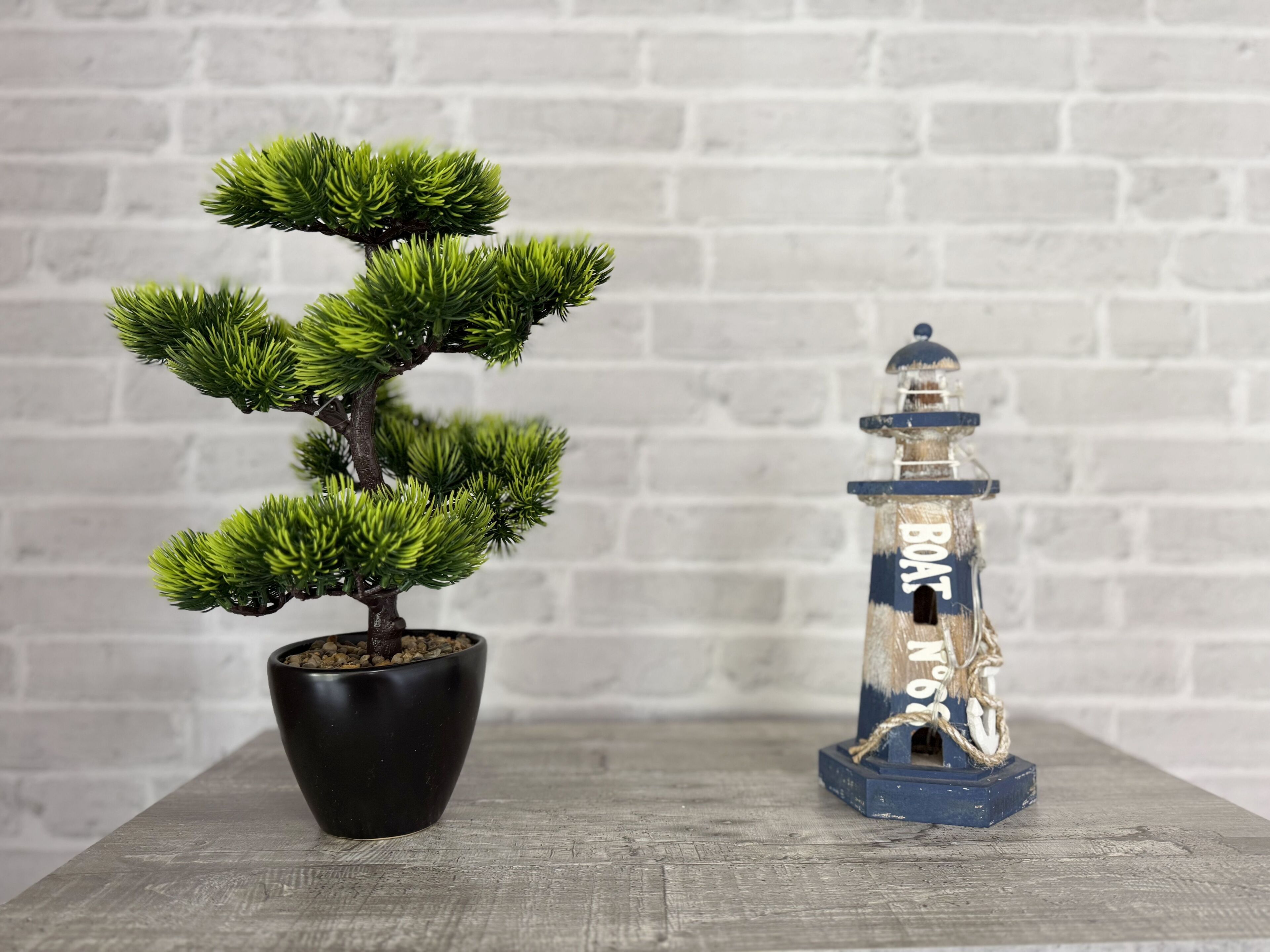 Un árbol bonsái en una maceta negra y un farol con forma de faro sobre una mesa de madera.