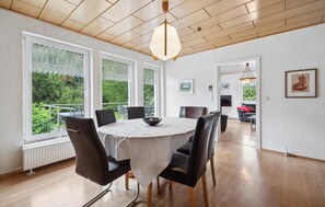 Een eethoek met een ronde tafel, stoelen en uitzicht op groen door grote ramen.