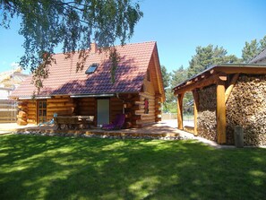 Une cabane en rondins avec un toit en tuiles, une terrasse avec une table et des chaises, et un espace abrité où est entreposé du bois de chauffage.
