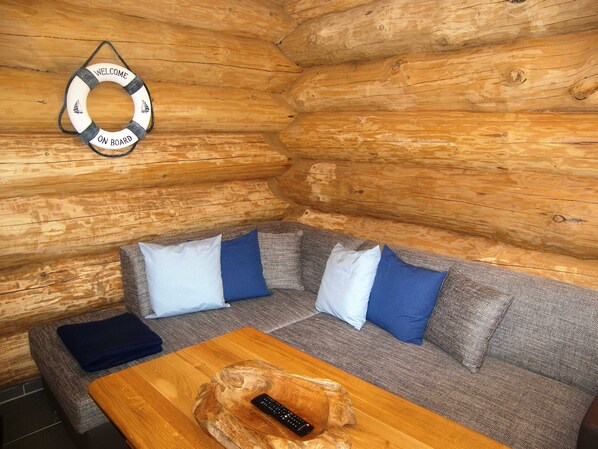 L’intérieur d’une cabane en rondins, avec un mur en bois, un canapé agrémenté de coussins bleus et blancs, et une table en bois sur laquelle repose une télécommande.