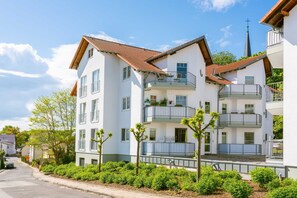 Un immeuble résidentiel à plusieurs étages doté de balcons, avec le clocher d’une église en arrière-plan et un ciel dégagé.