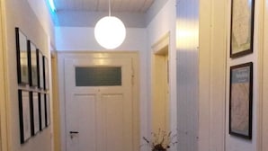 Un couloir avec une porte blanche au fond, un radiateur sur la droite et des tableaux encadrés accrochés aux murs.