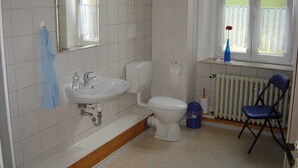 Une salle de bain avec un lavabo, des toilettes et un miroir, tous de couleur blanche. On y trouve également une chaise bleue et un radiateur près de la fenêtre.