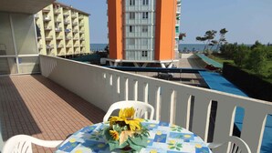 A balcony with a table covered in a tablecloth with lemon patterns, white chairs, and a flower arrangement. In the background, there are buildings and a view of the sea. - Holiday apartment 50 m to the beach (Porto Santa Margherita)