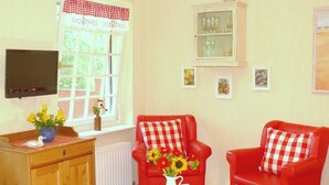 Una habitación con un sofá de cuero rojo, una mesa blanca, un aparador de madera, una ventana con cortina de cuadros rojos y un televisor montado.