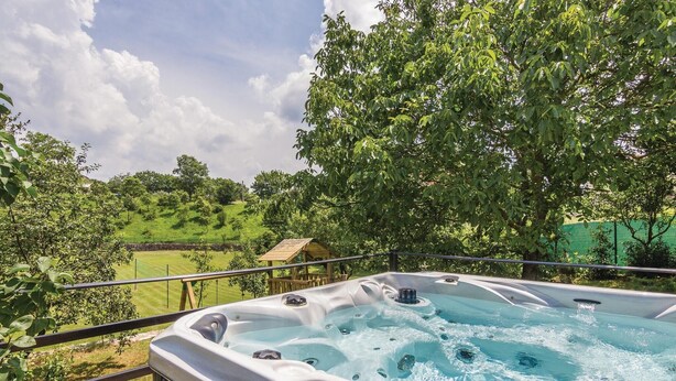 Een hottub met bedieningspaneel, omgeven door een houten terras en een metalen reling, met uitzicht op een gazon met bomen en een kleine houten constructie.