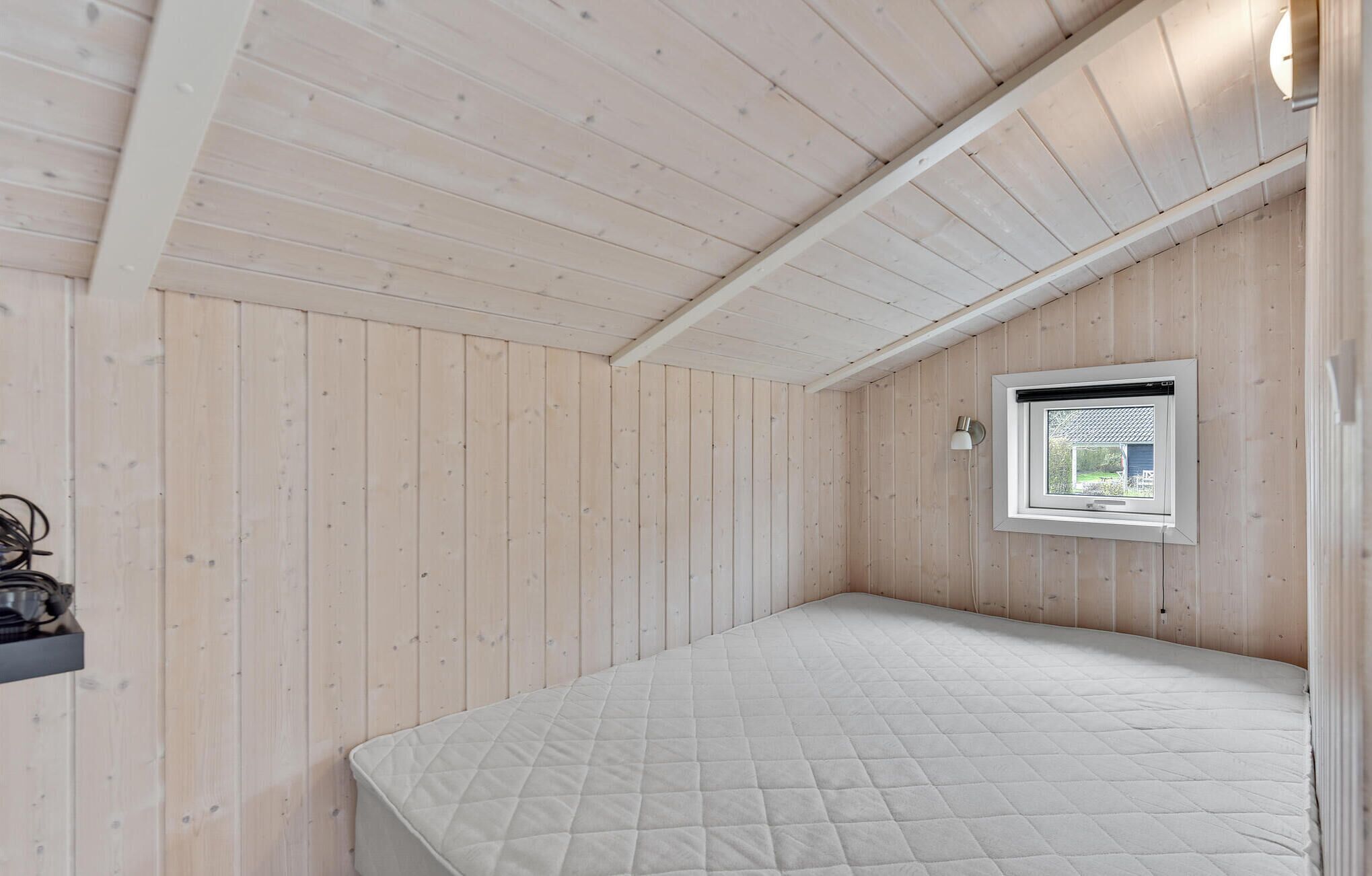Een kleine eenpersoonskamer met houten wanden en een schuine plafond. Er is een raam met uitzicht en een wandlamp.