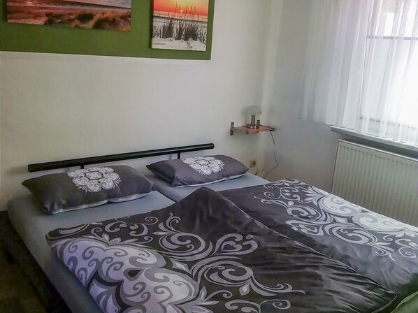 Un dormitorio con cama, almohadas, una mesita de noche, un estante en la pared y una ventana con cortinas.