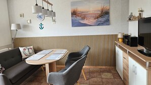 Una cocina moderna con comedor, que presenta una mesa de madera, sillas tapizadas de gris, una lámpara colgante y un cuadro de un atardecer sobre el agua en la pared.