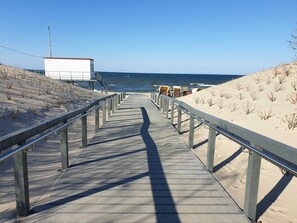 En træpromenade der fører til en strand med en hvid bygning og liggestole.