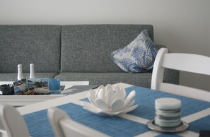 A living room with a grey sofa, a white chair, a table with a blue tablecloth, and a decorative white lotus flower centerpiece. - Haus Meeresblick FeWo Am Strand A 2.22 Ref. 128682 u 2 - FeWo Am Strand A 2.22 (Baabe)