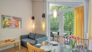 Een moderne woonkamer met een grijze bank, een houten eettafel gedekt voor twee personen, en een groot raam met gele gordijnen.