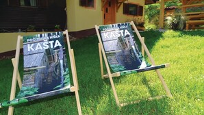 Twee schilderstatieven met posters van een rustiek houten huis en een stenen brug, staan op een gazon voor een gebouw met een houten hek en bloempotten.