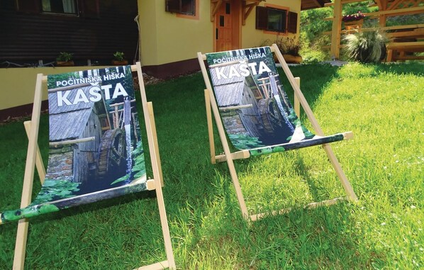Kaksi maalaustelinettä, joissa on maalauksia maalaismaisesta puutalosta ja kivisillasta. Ne on sijoitettu nurmikolle rakennuksen eteen, ja rakennuksella on puinen aita sekä ruukuissa olevia kasveja.