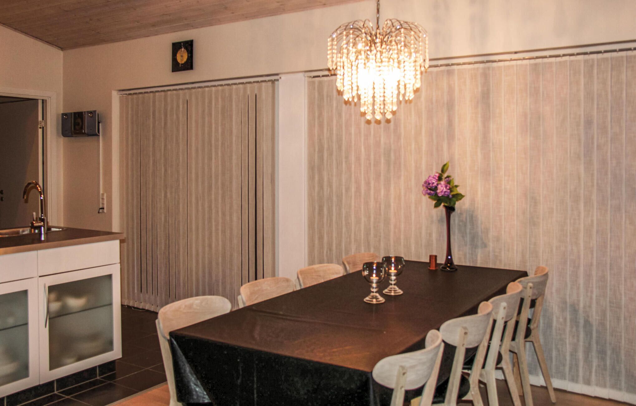 Un comedor con una mesa larga, sillas blancas, una lámpara de araña, un jarrón con flores y un reloj en la pared.