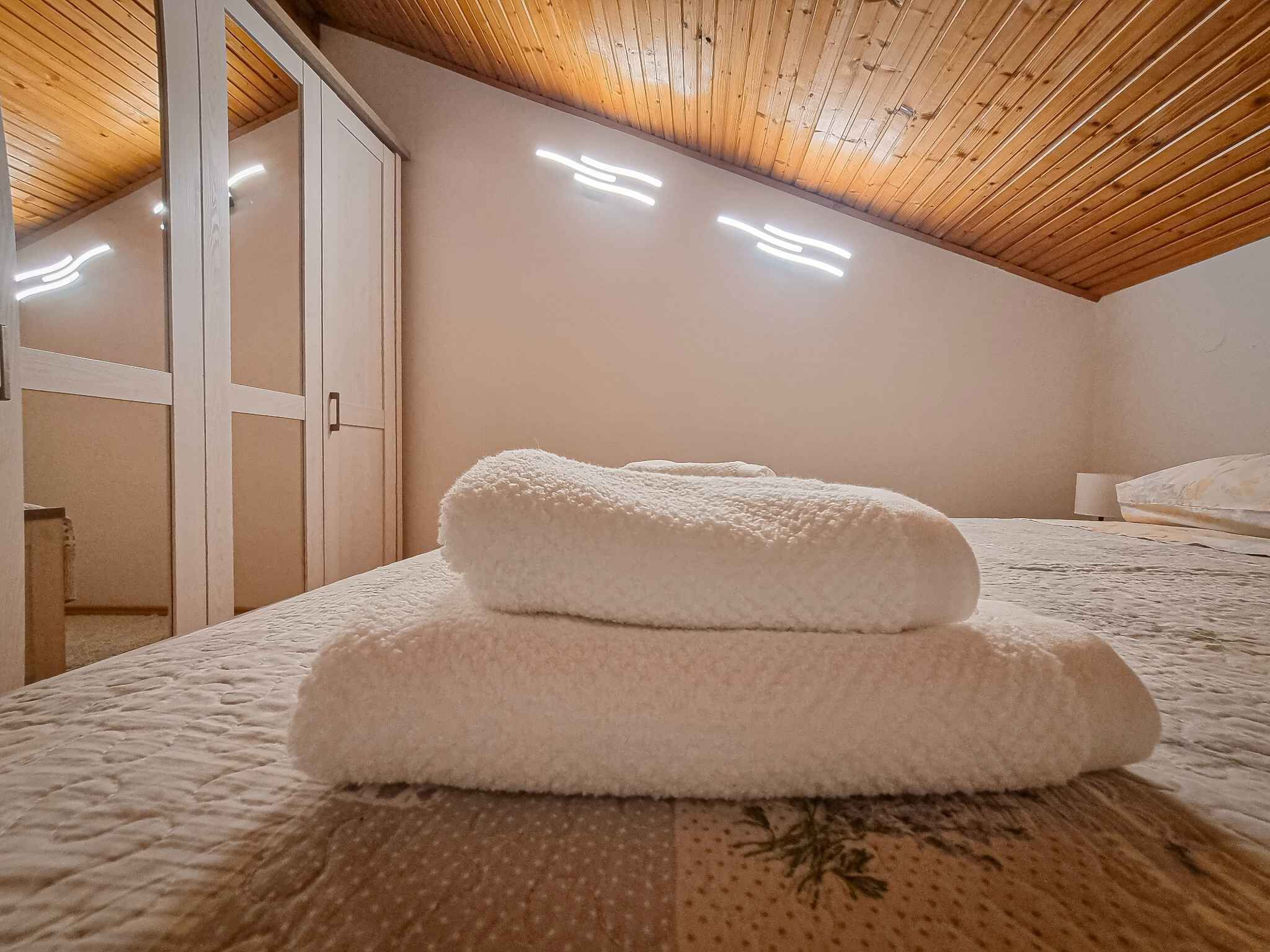 A bedroom with a wooden ceiling, a bed with a floral-patterned bedspread, and two rolled-up towels on the bed.