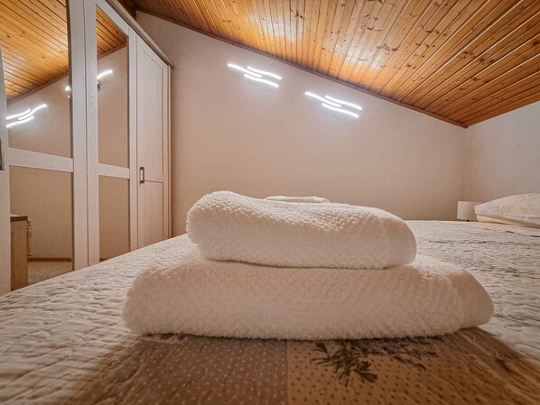 A bedroom with a wooden ceiling, a bed with a floral-patterned bedspread, and two rolled-up towels on the bed.
