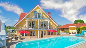 A two-story building with a swimming pool, outdoor seating, and a red-roofed structure nearby.