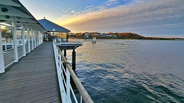 A waterfront pier with a covered seating area and a view of the water and distant buildings.