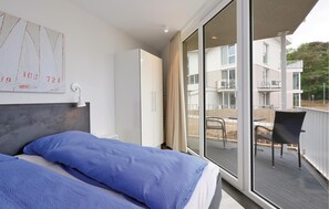 Een slaapkamer met een bed, een garderobe, een bureau en een groot raam met uitzicht op een balkon en gebouwen.