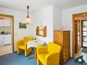 A compact room with a kitchen area, two yellow chairs, a small round table with a vase, a wooden cabinet, and a ceiling light fixture.