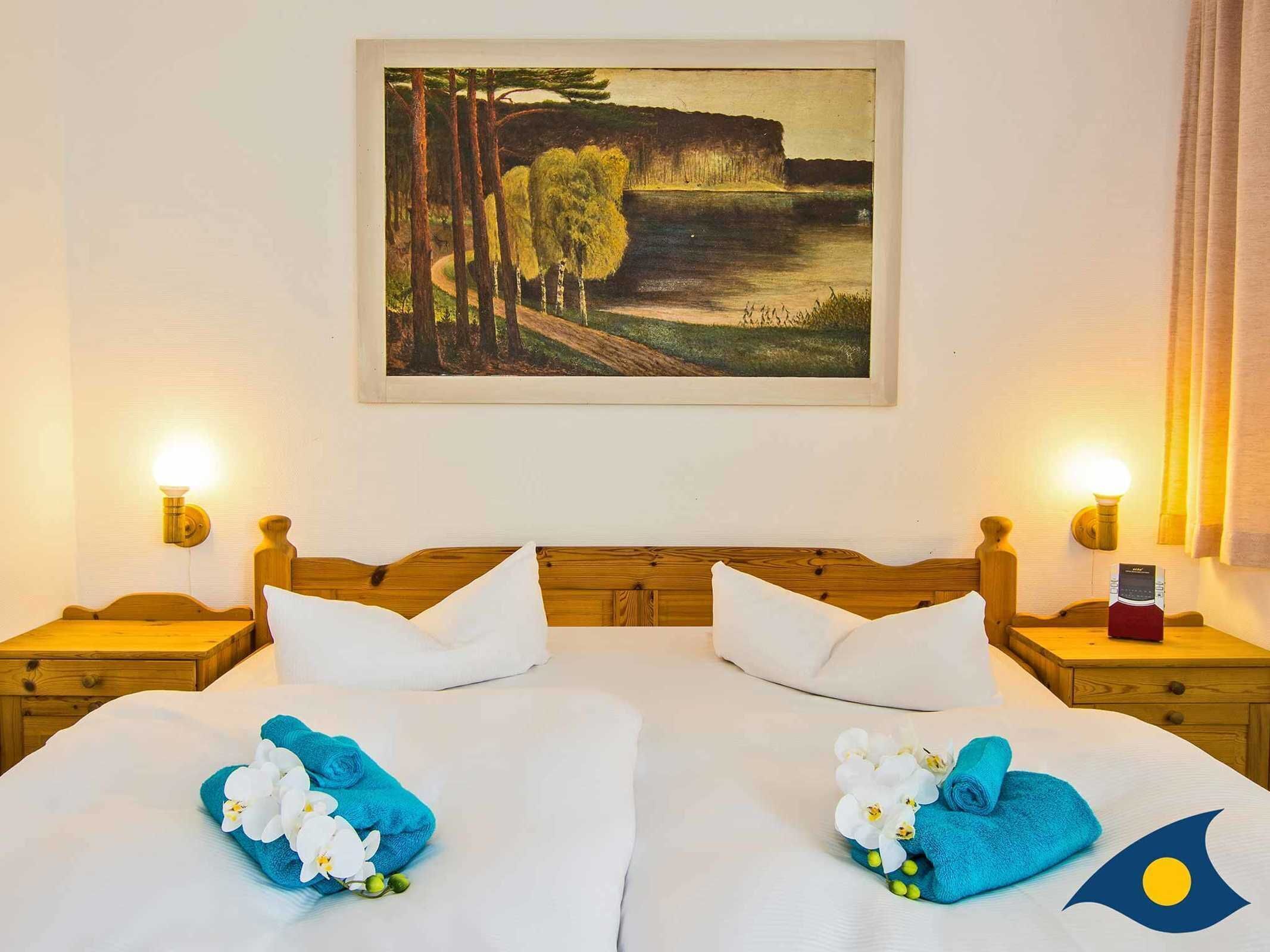 Ein Hotelzimmer mit zwei Betten, jeweils mit weißen Leintüchern und Handtüchern, die mit Blumen verziert sind. An der Wand hängt ein Landschaftsbild, und das Zimmer verfügt über hölzerne Kopfteile und Nachttische mit Lampen.