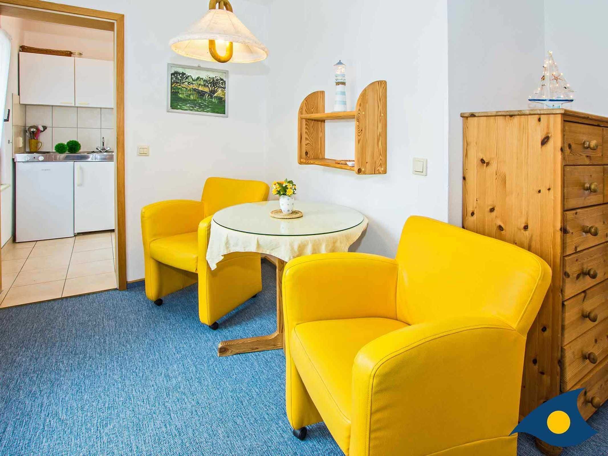 Ein Zimmer mit gelben Stühlen, einem kleinen runden Tisch, einem Holzschrank und einer Küchenzeile im Hintergrund.