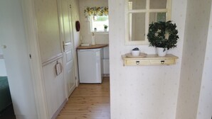 Un couloir étroit avec un plancher en bois, une porte blanche, un petit coin cuisine avec un réfrigérateur et une fenêtre ornée d’un rideau à motifs floraux.