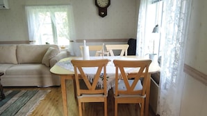 Un salon chaleureux avec un canapé beige, une table à manger en bois et deux chaises, une horloge au mur et des rideaux en dentelle aux fenêtres.