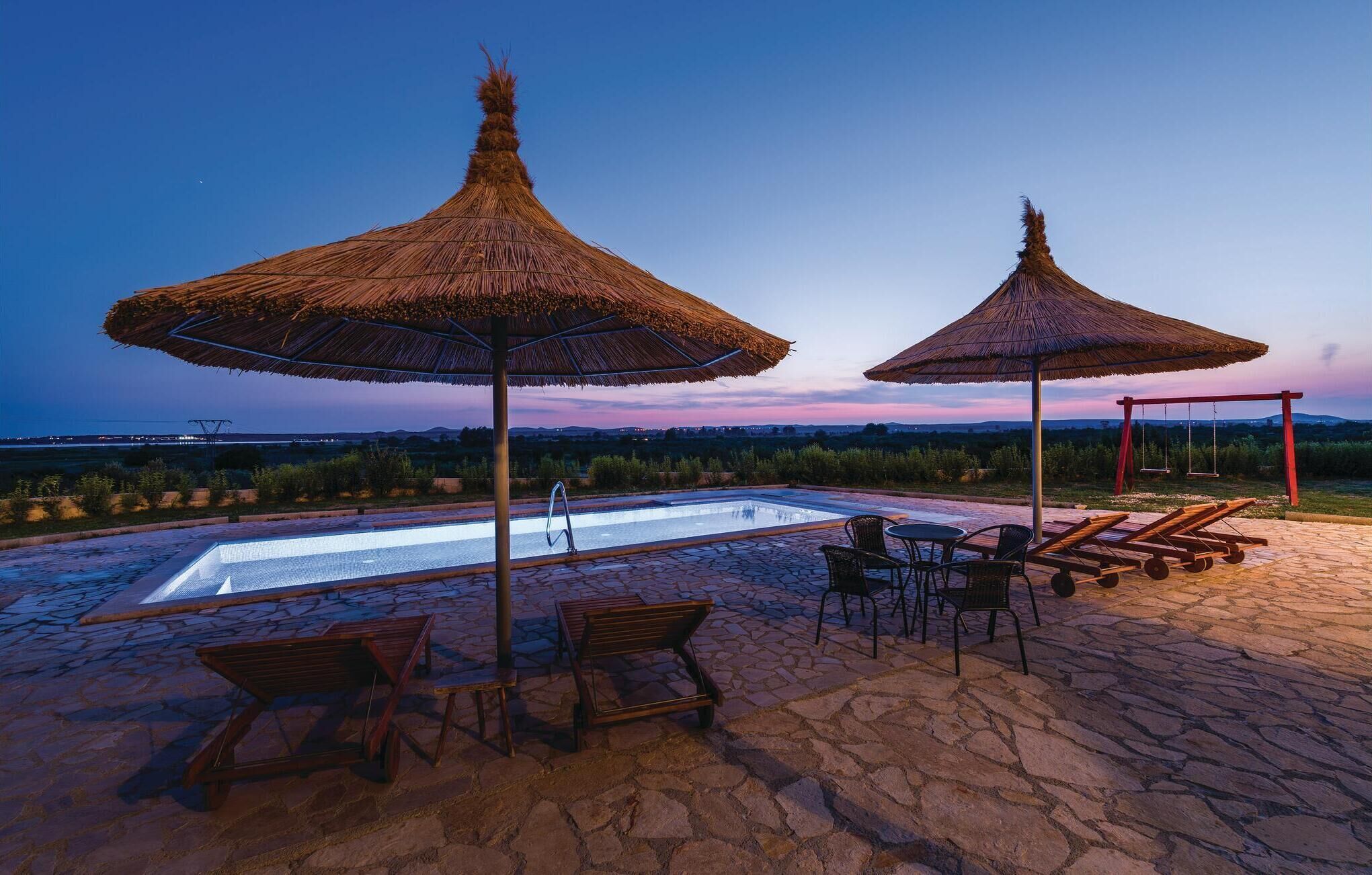A pool area with two thatched umbrellas, lounge chairs, and a stone patio.