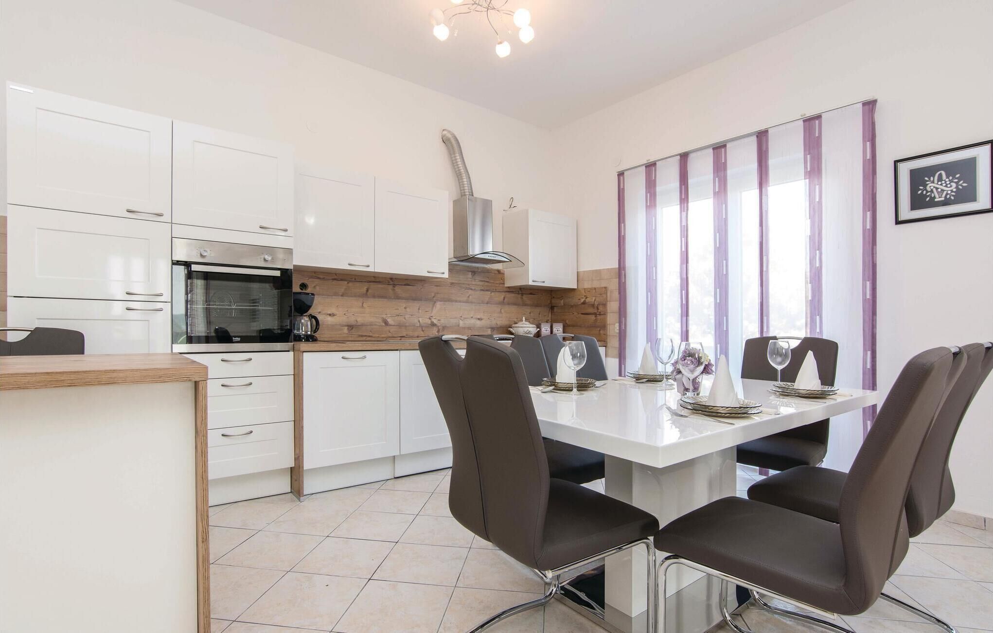 A modern kitchen with white cabinets, a built-in oven, and a coffee maker. There is a dining table with four chairs and a window with purple curtains.