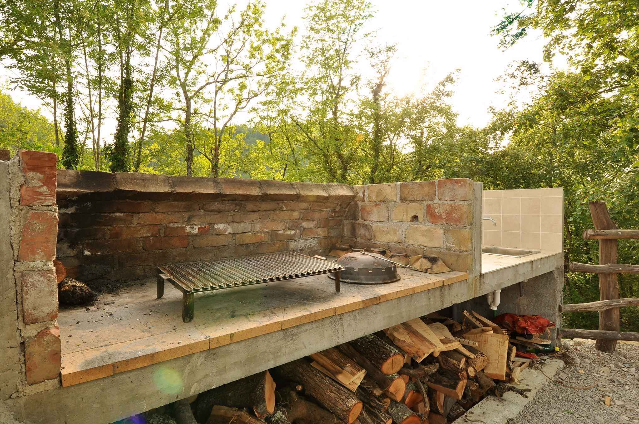 An outdoor brick oven with a metal grill and a sink, surrounded by stacked firewood.