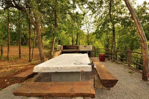 A rustic outdoor sleeping area with a wooden bench and a metal bed frame, surrounded by a wooden fence and trees.