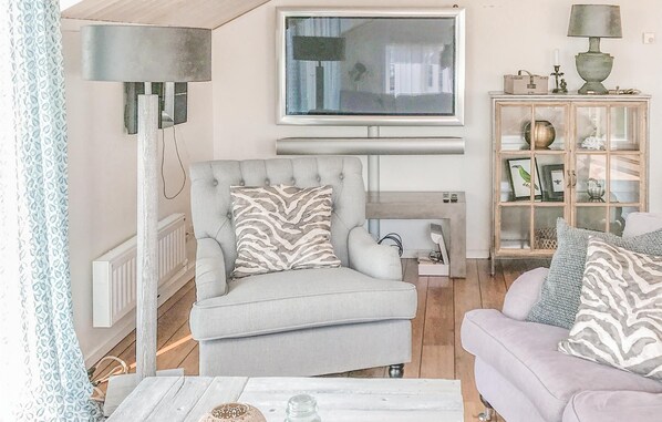 Un salon comprenant un fauteuil gris, un canapé blanc, un téléviseur fixé au mur, une lampe et un meuble avec des portes en verre.