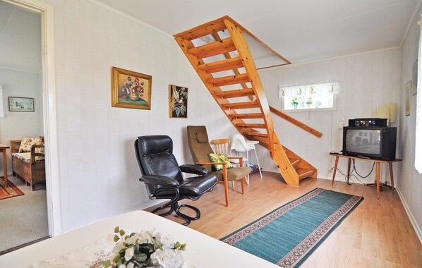 Un salon avec un escalier en bois, un fauteuil en cuir noir, une table en bois ornée d’un vase de fleurs, un téléviseur posé sur un meuble et une fenêtre avec des rideaux.