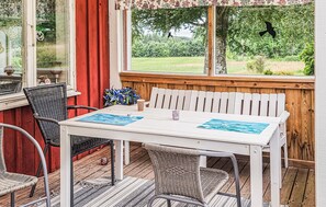 Un espace de restauration en plein air avec une table blanche et des chaises en osier, un rideau rouge et une vue sur la verdure par la fenêtre.