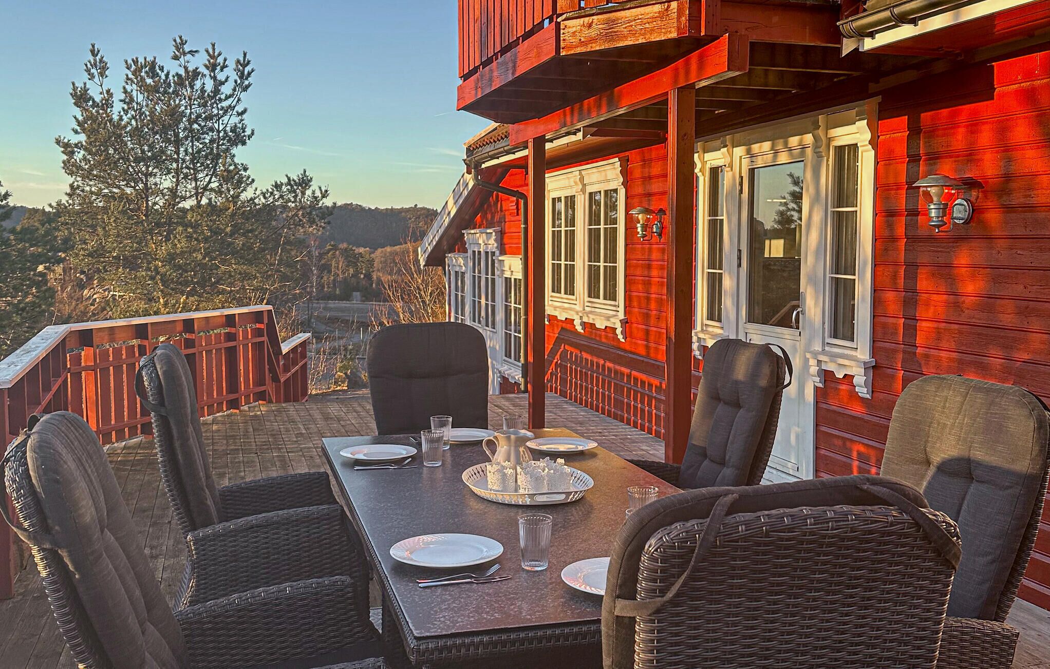 A dining area with a table set for four, surrounded by wicker chairs, on a wooden deck with a view of a red building and trees.