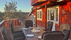 Ein Essbereich mit einem Tisch für vier Personen, umgeben von Korbstühlen, auf einer Holzterrasse mit Blick auf ein rotes Gebäude und Bäume.