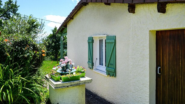 Una pequeña casa con fachada blanca, contraventanas verdes y puerta marrón. Un pequeño jardín con macetas y flores se encuentra al frente.