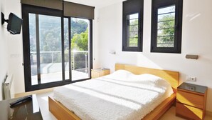 Un dormitorio con cama, mesitas de noche, televisor y ventanas amplias con vistas a zonas verdes.