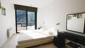 Un dormitorio con cama, escritorio, silla, radiador y un amplio ventanal con vista a árboles.