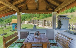 Un espace extérieur aménagé, abrité et doté d’une table en bois et de bancs, offrant une vue sur un paysage verdoyant, avec un mur de pierres et un petit bâtiment.