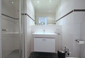 Un baño moderno con cabina de ducha de vidrio, un lavabo blanco con espejo encima y un cubo de basura negro.